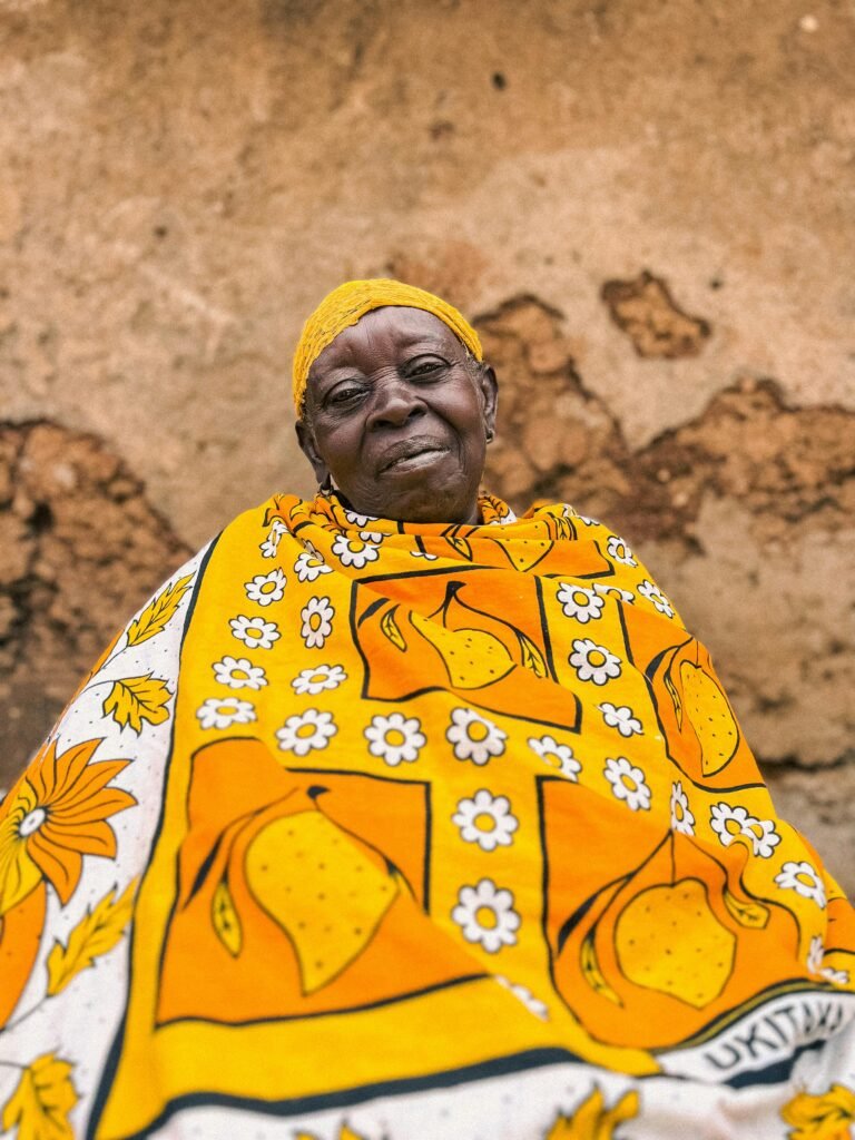Portrait of an elderly African woman in a vibrant yellow patterned wrap, smiling in front of a rustic wall.